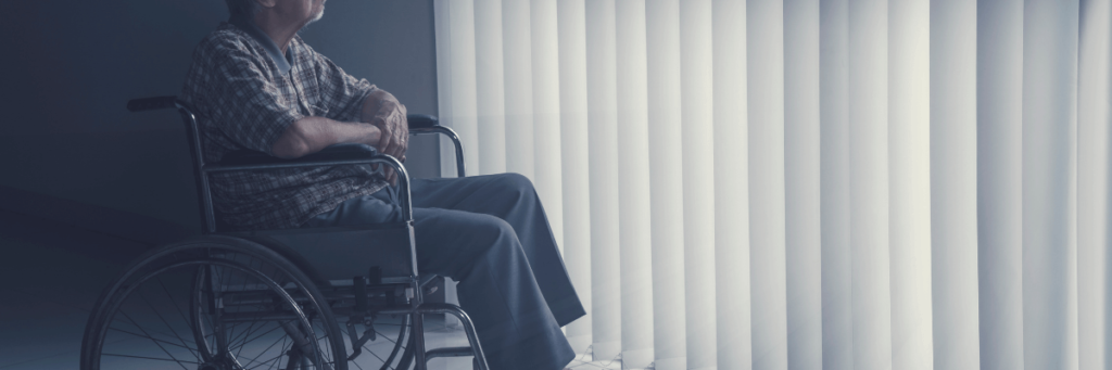 old person in a wheelchair staring out into a window. The room is dark and the curtains are pulled across. They look depressed.